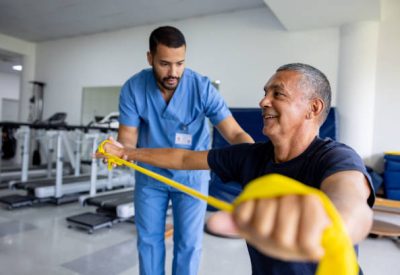 Mature Latin American man doing physical therapy exercises using a stretch band with the assistance of his therapist